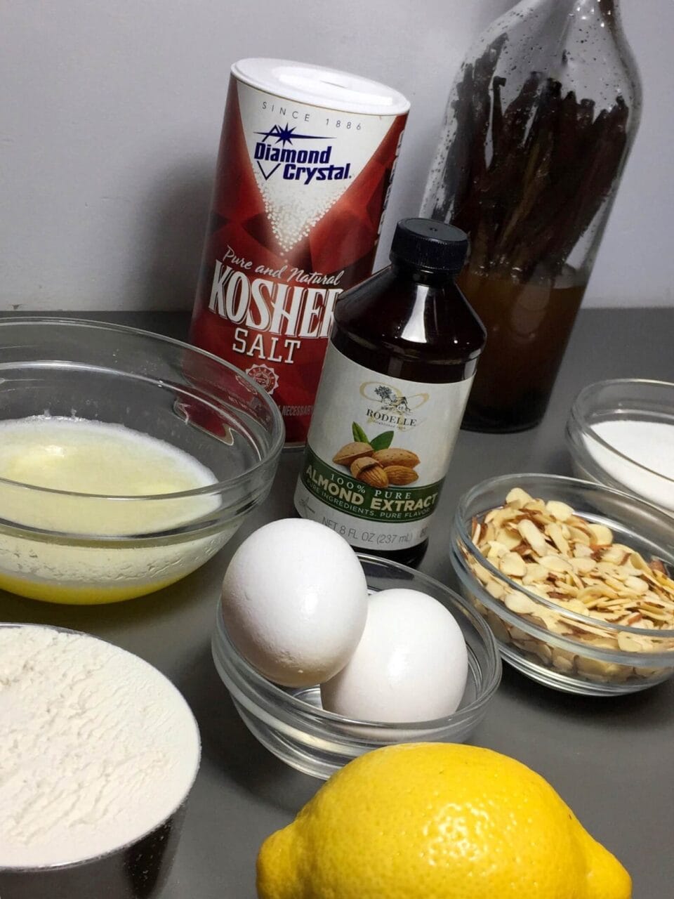 Ingredients for baking include kosher salt, almond extract, eggs, lemon, flour, melted butter, sliced almonds, and vanilla beans.