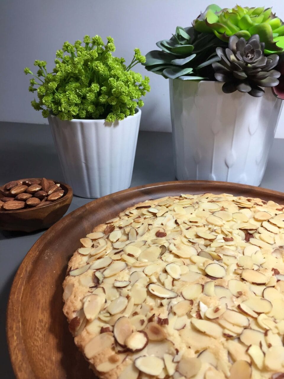 Almond cake on a wooden tray with decorative succulents and a bowl of almonds in the background.