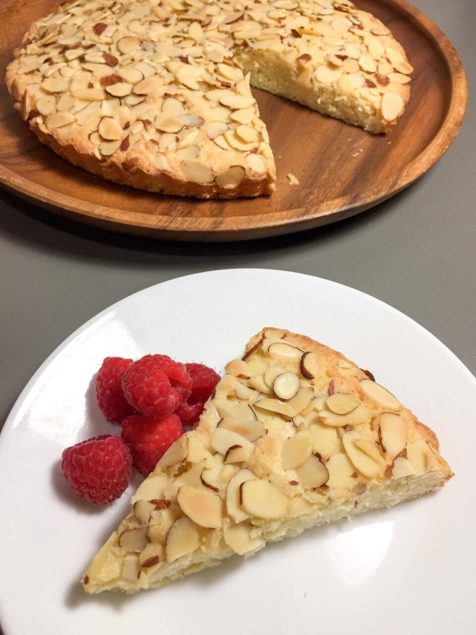 A slice of almond cake served with fresh raspberries on a white plate, with the remaining cake on a wooden tray in the background.