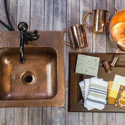A copper sink and faucet set on a rustic wooden surface, accompanied by copper mugs, a tray with fabric swatches, and kitchen accessories, creating a warm and earthy kitchen design aesthetic.
