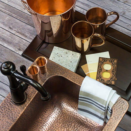 A copper sink with a dark faucet is accompanied by copper mugs, a small bucket, fabric swatches, and design samples, all set on a rustic wooden surface, showcasing a warm and coordinated kitchen design theme.