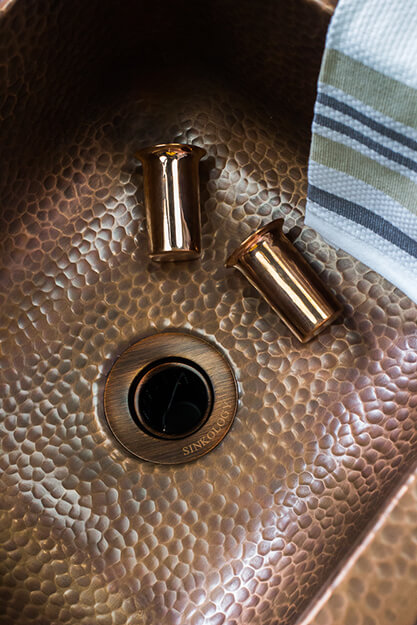 A close-up of a hammered copper sink with two copper pieces and a striped cloth draped over the edge, highlighting the rich texture and warmth of the materials.