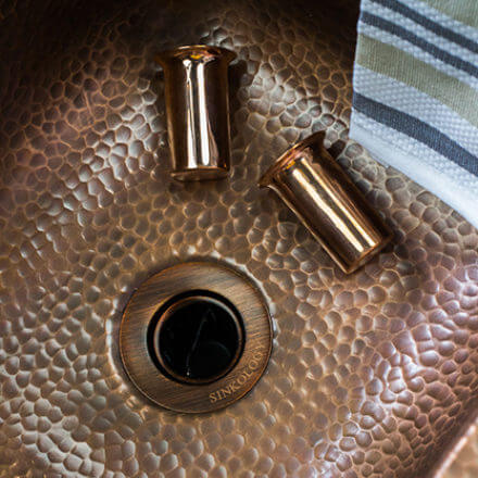 A close-up of a hammered copper sink with two copper pieces and a striped cloth draped over the edge, highlighting the rich texture and warmth of the materials.