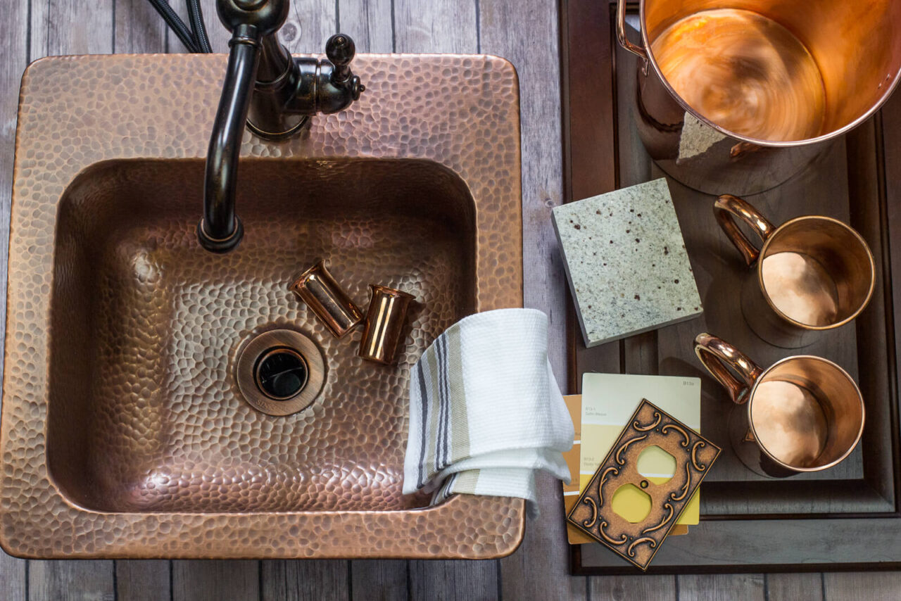A top view of a hammered copper sink with a dark faucet, copper accessories, fabric swatches, and design samples laid out on a rustic wooden surface, creating a coordinated kitchen design setup.