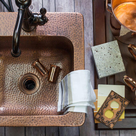 A top-down view of a hammered copper sink with a dark faucet, accompanied by copper mugs, fabric swatches, and design samples, all arranged on a rustic wooden surface.