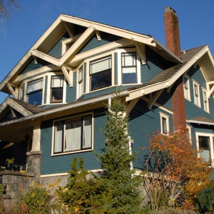 A classic Craftsman-style house with blue siding, white trim, and a prominent brick chimney, surrounded by a well-maintained garden and trees.